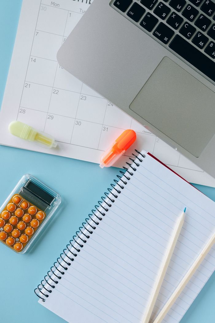 Flat lay of a workspace with a laptop, notebook, calculator, and calendar on a blue background.